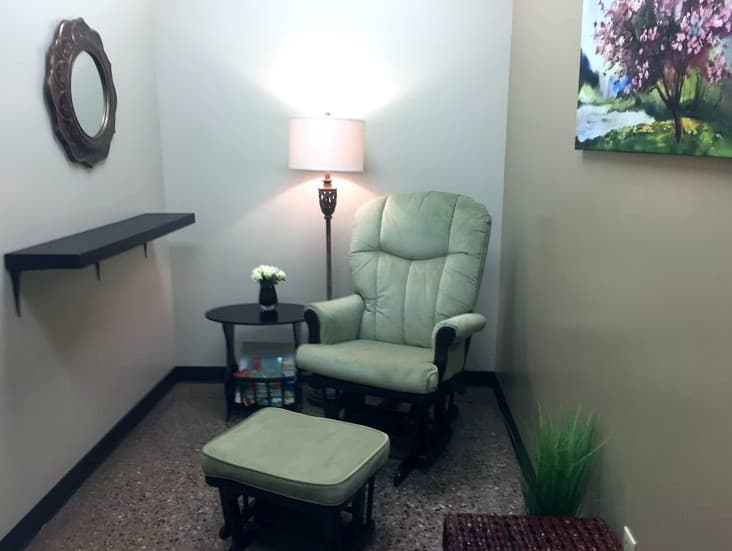 Interior view of a MomDoc Charities Mother's Room with a glider chair, ottoman, warm lamp, and children's books